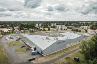 Aerial photo of the completed daycare: a long, low building with grey standing-seam metal roofs and cladding, beside a road and open fields.