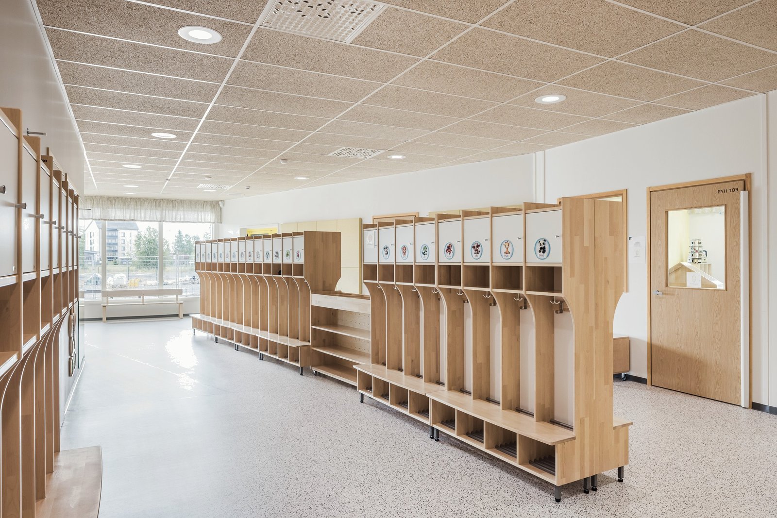 Interior cloakroom with rows of light birch cubbies and coat hooks for children, under a white acoustic ceiling.