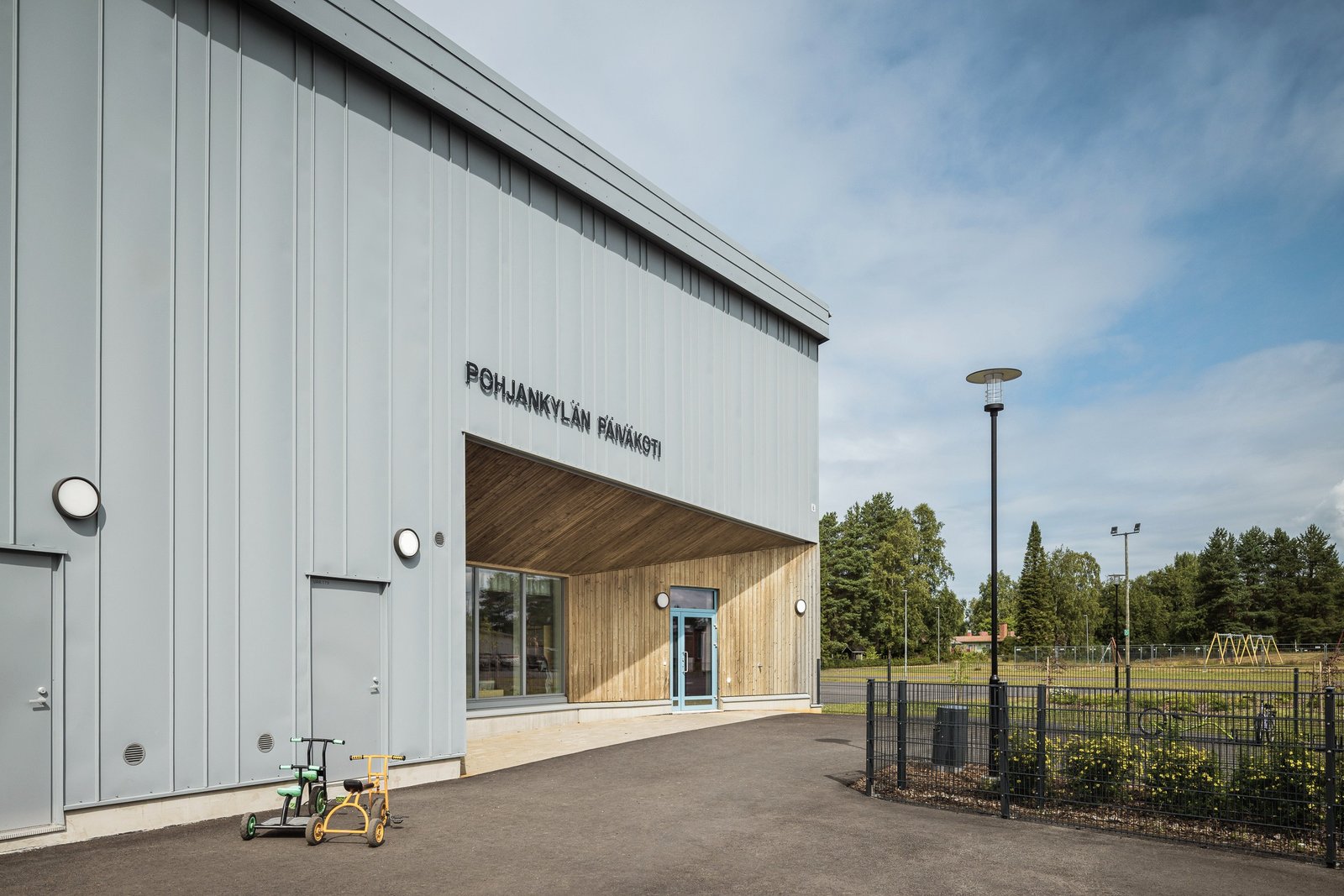 Main entrance: grey vertical cladding cut back beneath a warm timber-lined canopy, with the sign Pohjankylän päiväkoti.