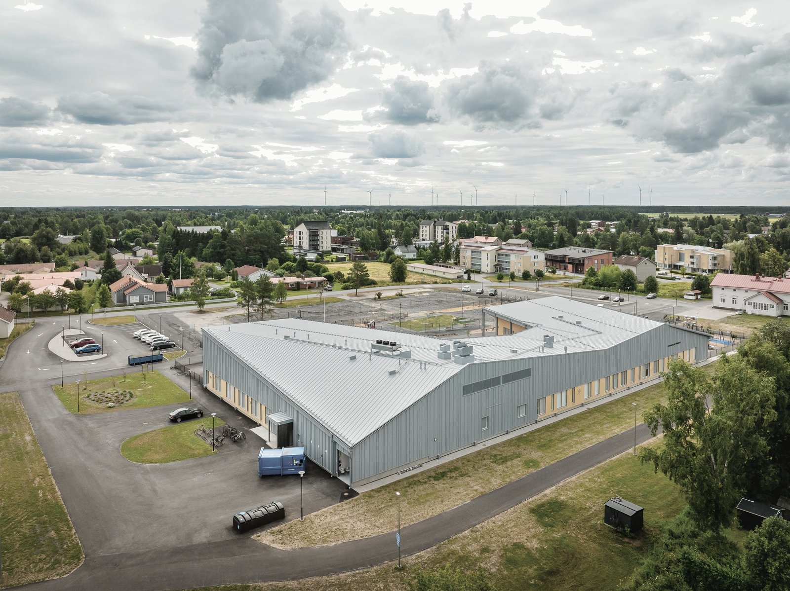 Aerial photo of the completed daycare: a long, low building with grey standing-seam metal roofs and cladding, beside a road and open fields.