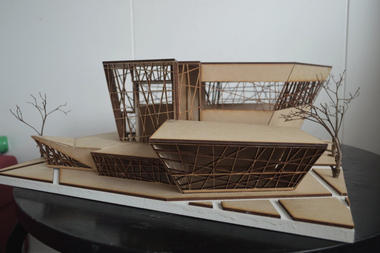 Study model in laser-cut board seen from the front: low angular volumes with cut lattice façades beneath a tall cantilevered gallery block, with twig trees on a paved base.